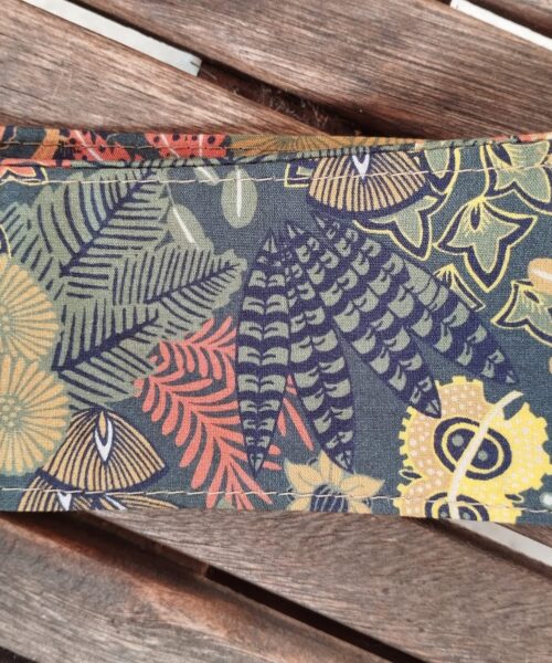 Gros plan du Bandeau Simple, montrant son motif botanique coloré avec des feuilles, des fougères et des fleurs dans des tons jaunes, verts, orange et bleus, affichés sur une surface en bois avec des planches visibles.